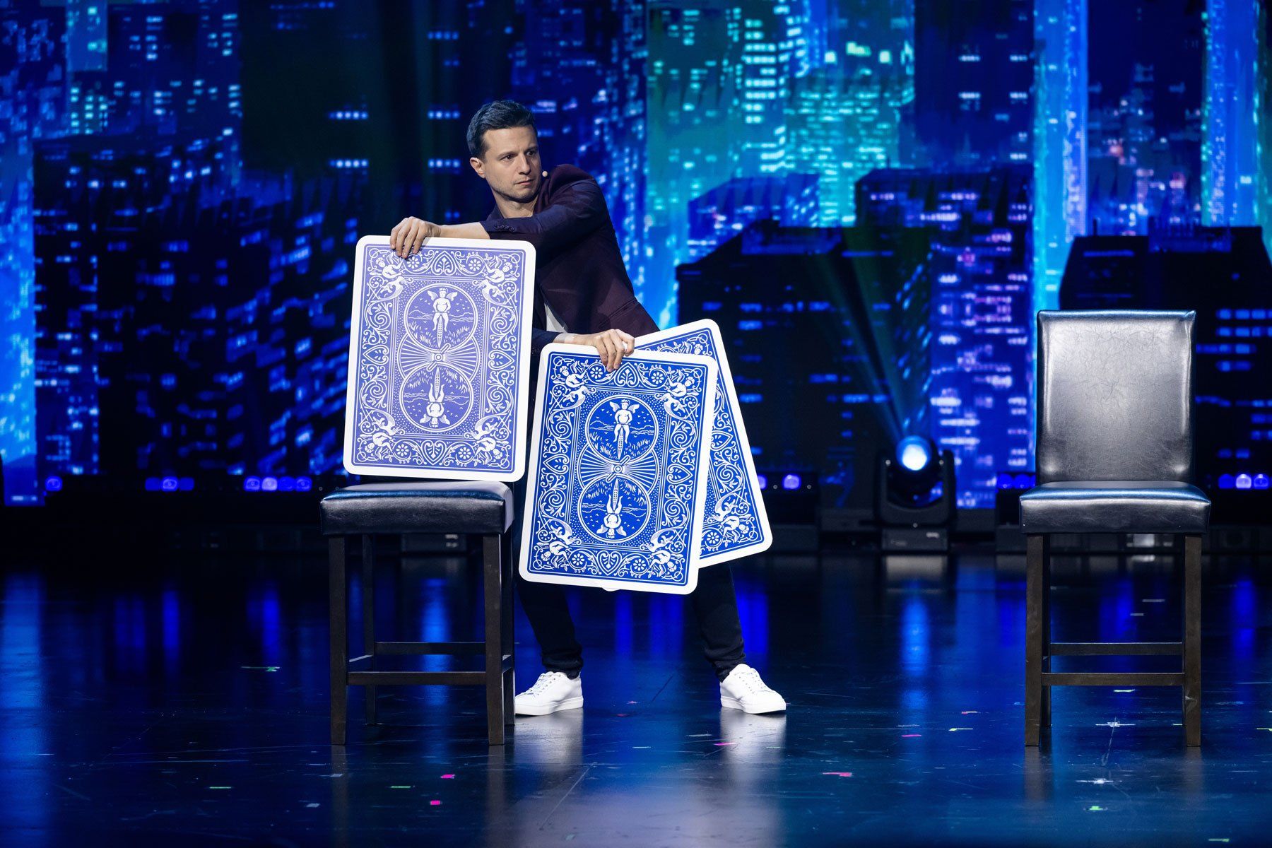 Magician on a theater stage performing with oversized blue playing cards between two stools against an illuminated city skyline backdrop.