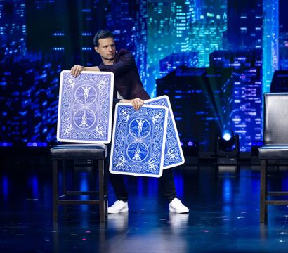 Magician on a theater stage performing with oversized blue playing cards between two stools against an illuminated city skyline backdrop.