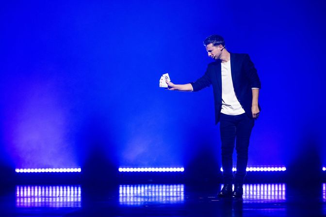 Magician performing on a blue-lit stage, extending a fan of playing cards toward the audience with row lights glowing behind him