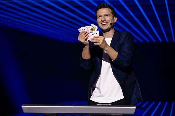 Smiling stage magician performing a card trick with a headset mic, fanned playing cards and a table under blue geometric stage lights at a live show.