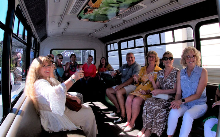 Smiling group of adults seated inside a daytime party shuttle, holding drinks and toasting while large windows reveal a city street outside.