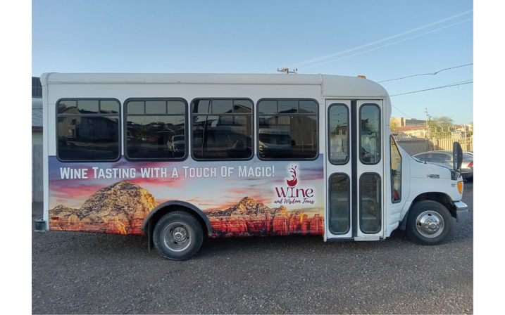 White shuttle bus parked on gravel, wrapped in a colorful wine-tasting mural of red-rock cliffs at sunset with the slogan Wine Tasting with a Touch of Magic! under a clear blue sky.