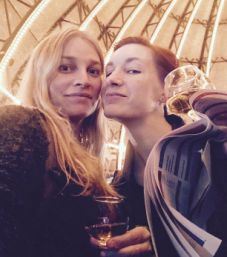 Two people smiling under a glowing canopy of arched lights, holding wine glasses and a folded event program — cozy festive night out.