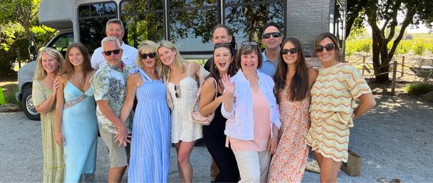 Smiling group of friends and family in summer dresses and casual wear posing in front of a shuttle at a sunny vineyard wine-country tasting stop with trees and grape rows in the background