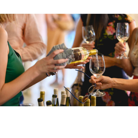 Close-up of white wine being poured into a glass at a lively wine-tasting event, with wine bottles and glasses on the table