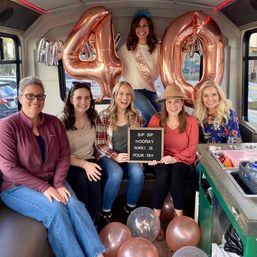 Six women smiling inside a party bus celebrating a 40th birthday with large rose-gold '40' balloons, a birthday sash, a playful letterboard message, and scattered rose-gold balloons.