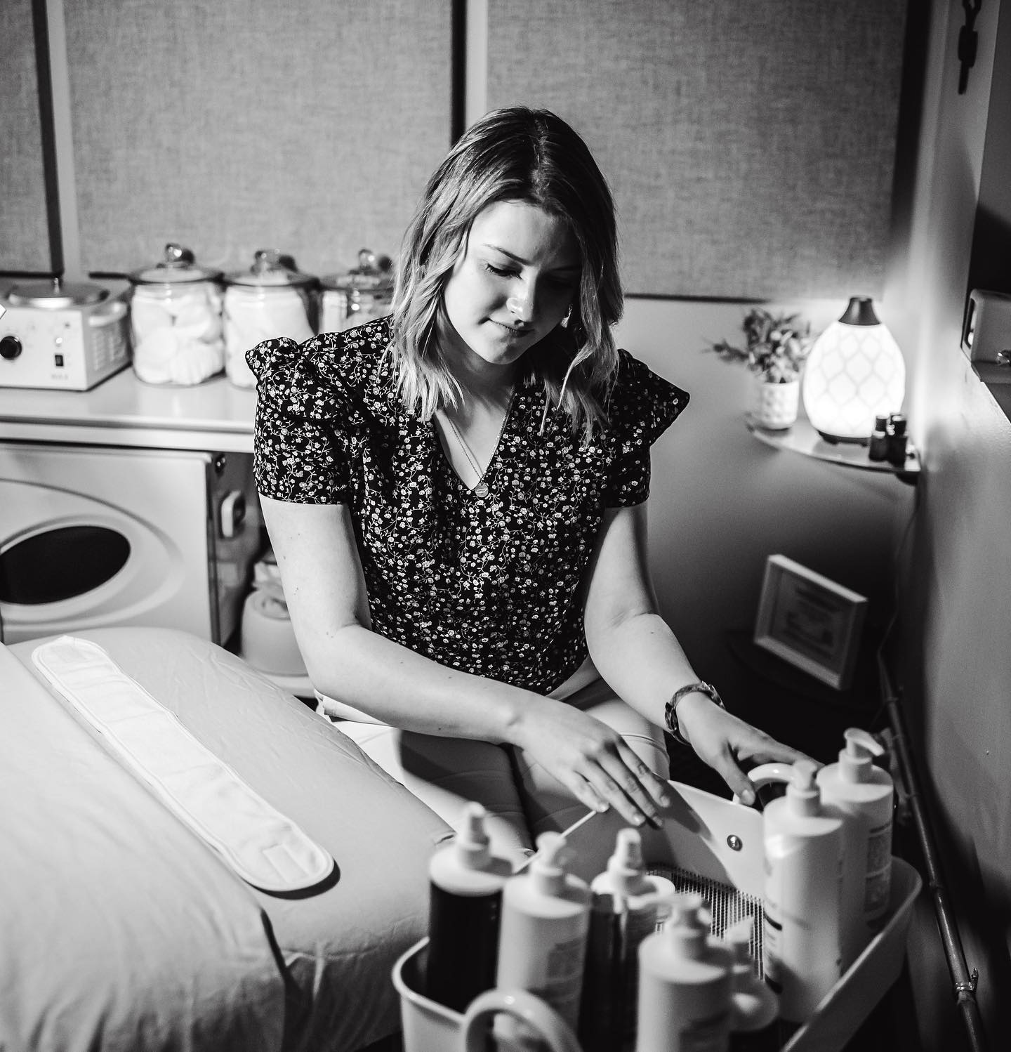 Cozy spa treatment room with an esthetician arranging lotion and oil bottles on a trolley beside a prepared massage table, jars of supplies and a glowing diffuser in the background.