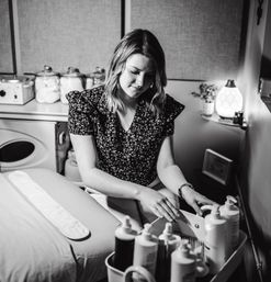Cozy spa treatment room with an esthetician arranging lotion and oil bottles on a trolley beside a prepared massage table, jars of supplies and a glowing diffuser in the background.