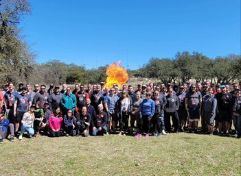 Outdoor team-building group photo on a grassy field with dozens of people posing in front of a bright orange fireball demonstration, clear blue sky and oak trees in the background