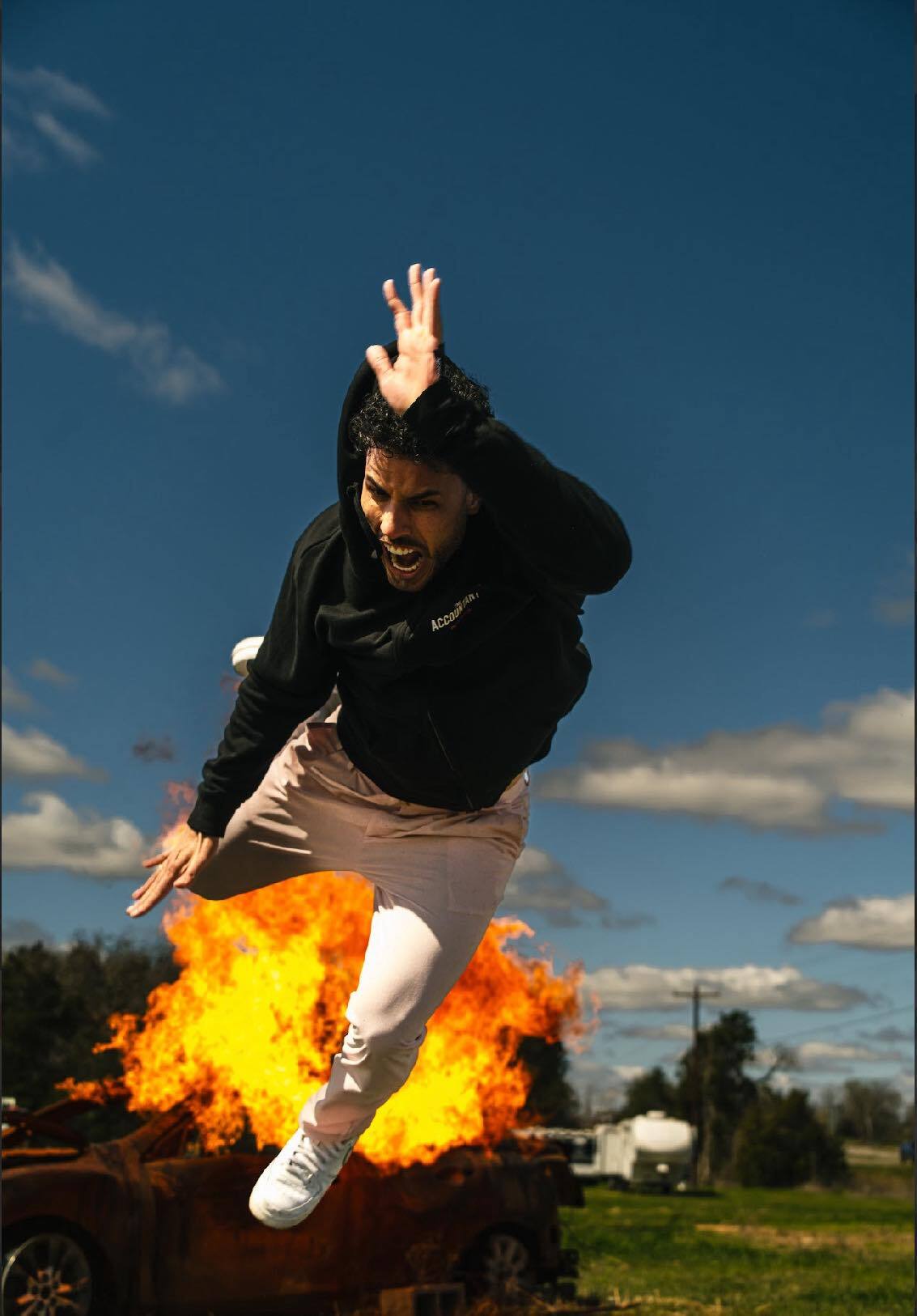 Person in a black hoodie leaping dramatically away from a fiery car explosion in an open field under a vivid blue sky — outdoor action scene