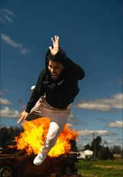 Person in a black hoodie leaping dramatically away from a fiery car explosion in an open field under a vivid blue sky — outdoor action scene