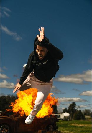 Person in a black hoodie leaping dramatically away from a fiery car explosion in an open field under a vivid blue sky — outdoor action scene