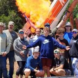 Group of teens and adults posing outdoors as a small airplane fuselage erupts in a large fireball behind them, several people smiling and holding hammers