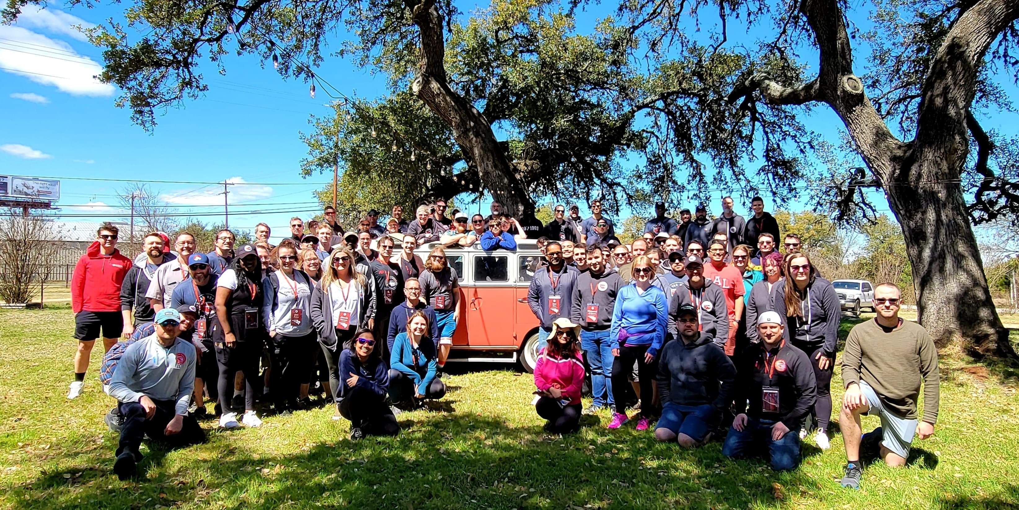 Large outdoor team photo of smiling people posing around a red vintage Volkswagen van on a sunny day beneath sprawling oak trees in a grassy park.