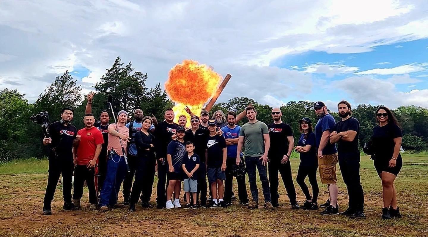 Large mixed-age film crew and friends posing in a grassy rural field with camera gear and kids, as a dramatic orange explosion/fireball erupts behind them against a partly cloudy sky — outdoor stunt/film shoot scene.