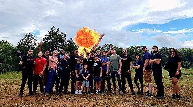 Large mixed-age film crew and friends posing in a grassy rural field with camera gear and kids, as a dramatic orange explosion/fireball erupts behind them against a partly cloudy sky — outdoor stunt/film shoot scene.