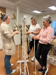 Three people in a cozy craft studio braiding macramé plant hangers on a standing frame, with wooden floors, woven pendant lights, and shelves of home goods in the background.