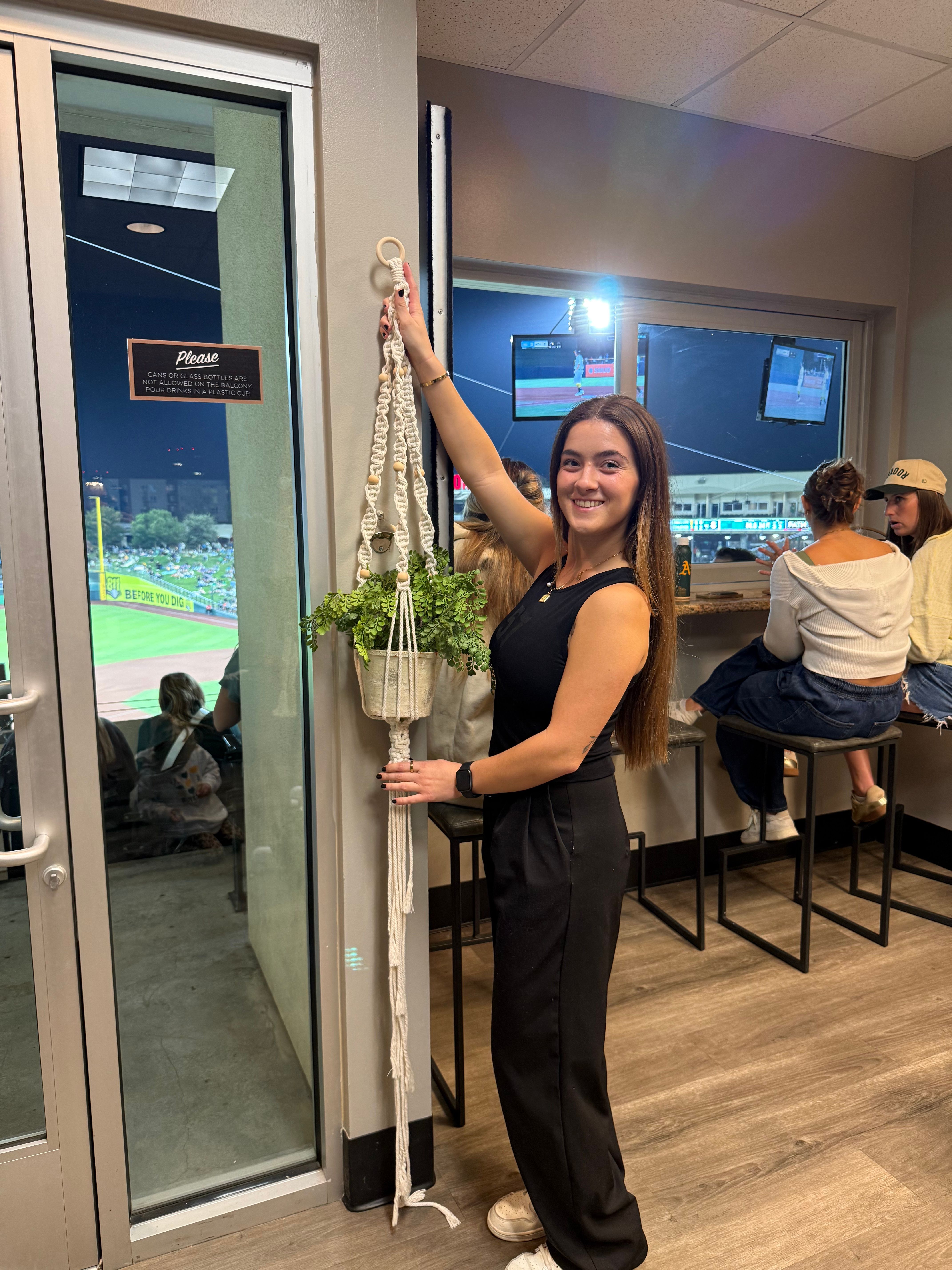 Smiling woman in a stadium suite holds a long macramé plant hanger with a green potted plant while a baseball game plays outside
