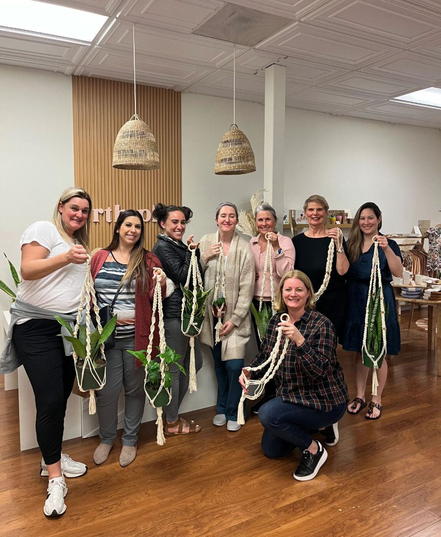 Group of eight women in a cozy craft studio proudly holding handmade macramé plant hangers with potted green plants under woven pendant lights