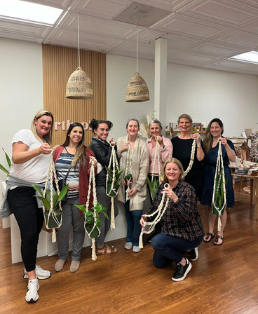 Group of eight women in a cozy craft studio proudly holding handmade macramé plant hangers with potted green plants under woven pendant lights