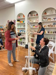 Women crafting macramé plant hangers in a cozy boutique workshop, laughing and knotting cotton cords among shelves of skincare and home goods on wood floors.