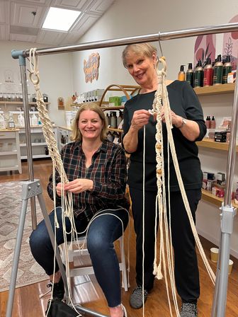 Two smiling women in a cozy craft studio weaving macramé plant hangers on a metal frame, wooden beads and product-lined shelves in the background.