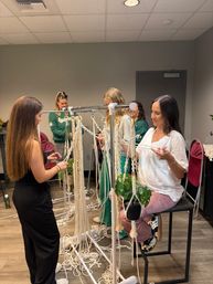 Women at an indoor macramé workshop crafting plant hangers from cotton cords on a metal rack with a potted plant.