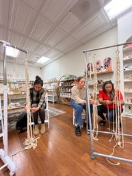 Three people crafting macramé plant hangers in a bright boutique craft studio, threading cotton cords on metal racks amid wooden floors, shelves of craft supplies and cozy retail displays.