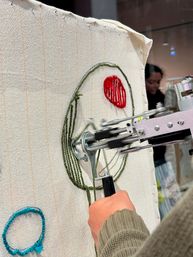 Close-up of a hand in a green ribbed sweater operating a small embroidery machine stitching a green circular outline and a red textured oval onto cream woven fabric, with a teal stitched loop and blurred background.