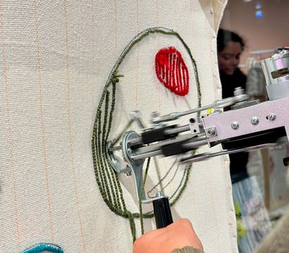 Close-up of a hand in a green ribbed sweater operating a small embroidery machine stitching a green circular outline and a red textured oval onto cream woven fabric, with a teal stitched loop and blurred background.