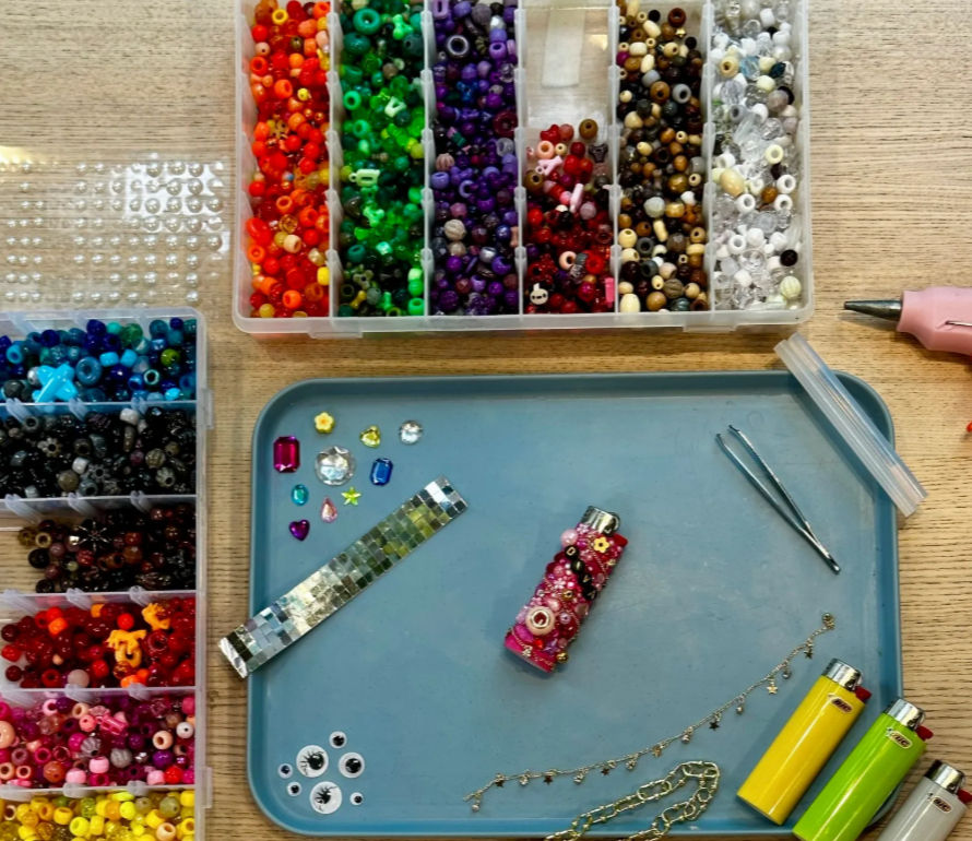 Colorful DIY jewelry-making workspace on a light wood table: organized plastic trays of multicolored beads, rhinestones and googly eyes, a blue work tray with tweezers, mosaic strip, decorated lighter, chain and three bright lighters — craft supplies for beading and decoration.