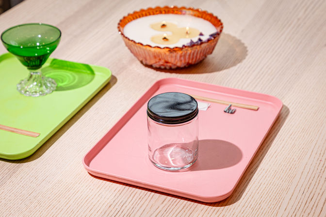 Clear glass jar with black lid centered on a pink serving tray on a light wood table, nearby green dessert glass on a lime tray and an amber scalloped candle bowl in the background — bright tabletop display.