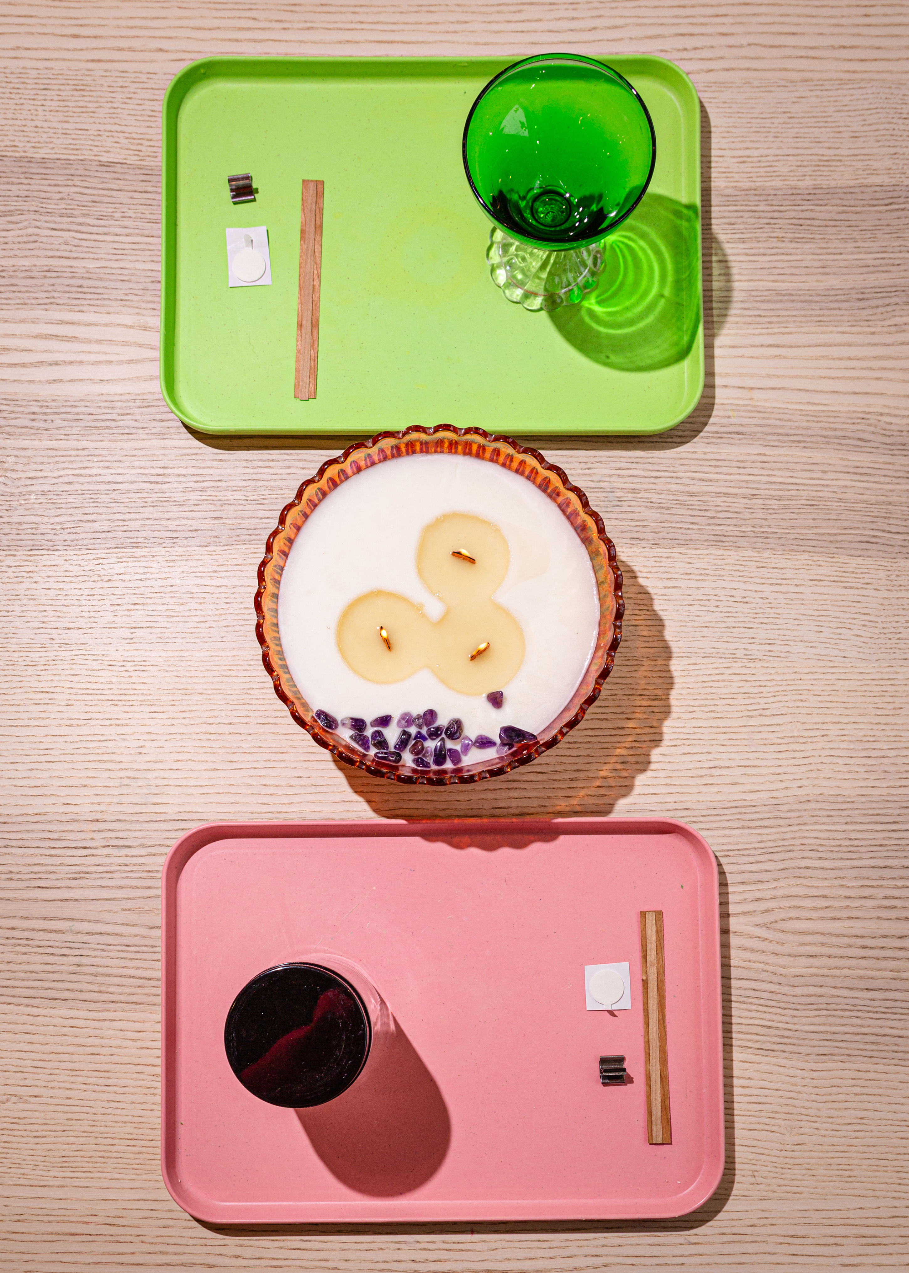 Top-down view of a light wood tabletop styled with a green tray holding a green goblet, a pink tray with a black cup and small accessories, and a scalloped glass candle bowl with three wicks and purple crystal chips — minimalist tabletop decor.