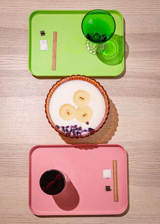 Top-down view of a light wood tabletop styled with a green tray holding a green goblet, a pink tray with a black cup and small accessories, and a scalloped glass candle bowl with three wicks and purple crystal chips — minimalist tabletop decor.