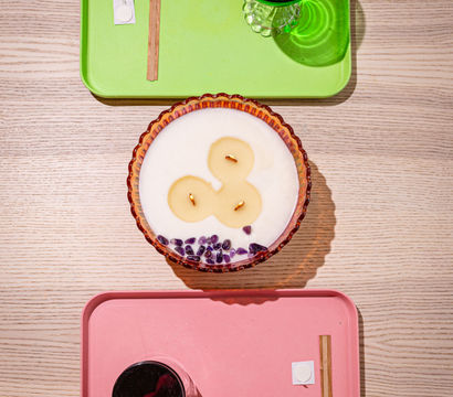 Top-down view of a light wood tabletop styled with a green tray holding a green goblet, a pink tray with a black cup and small accessories, and a scalloped glass candle bowl with three wicks and purple crystal chips — minimalist tabletop decor.