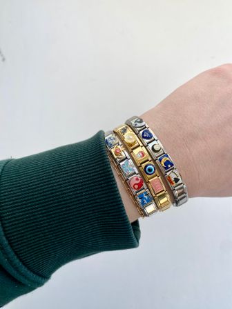 Close-up of a wrist wearing three stacked silver and gold Italian charm bracelets with colorful enamel charms (heart, yin-yang, evil eye, butterfly, pearl) against a white background and dark green ribbed sleeve, fashion accessory shot.