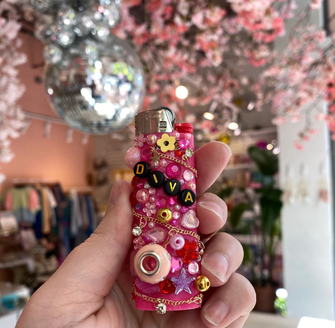 Hand holding a sparkly pink lighter covered in beads, gems, chains and black letter beads spelling DIVA, with a blurred boutique interior, disco ball and pink blossom decor in the background.