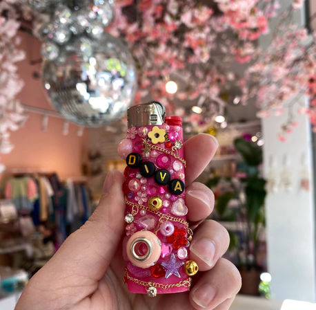 Hand holding a sparkly pink lighter covered in beads, gems, chains and black letter beads spelling DIVA, with a blurred boutique interior, disco ball and pink blossom decor in the background.