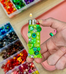 Sparkling lime-green lighter decorated with green rhinestones, beads and letter beads spelling "SLAY," held over a craft table with compartmented rainbow bead organizers