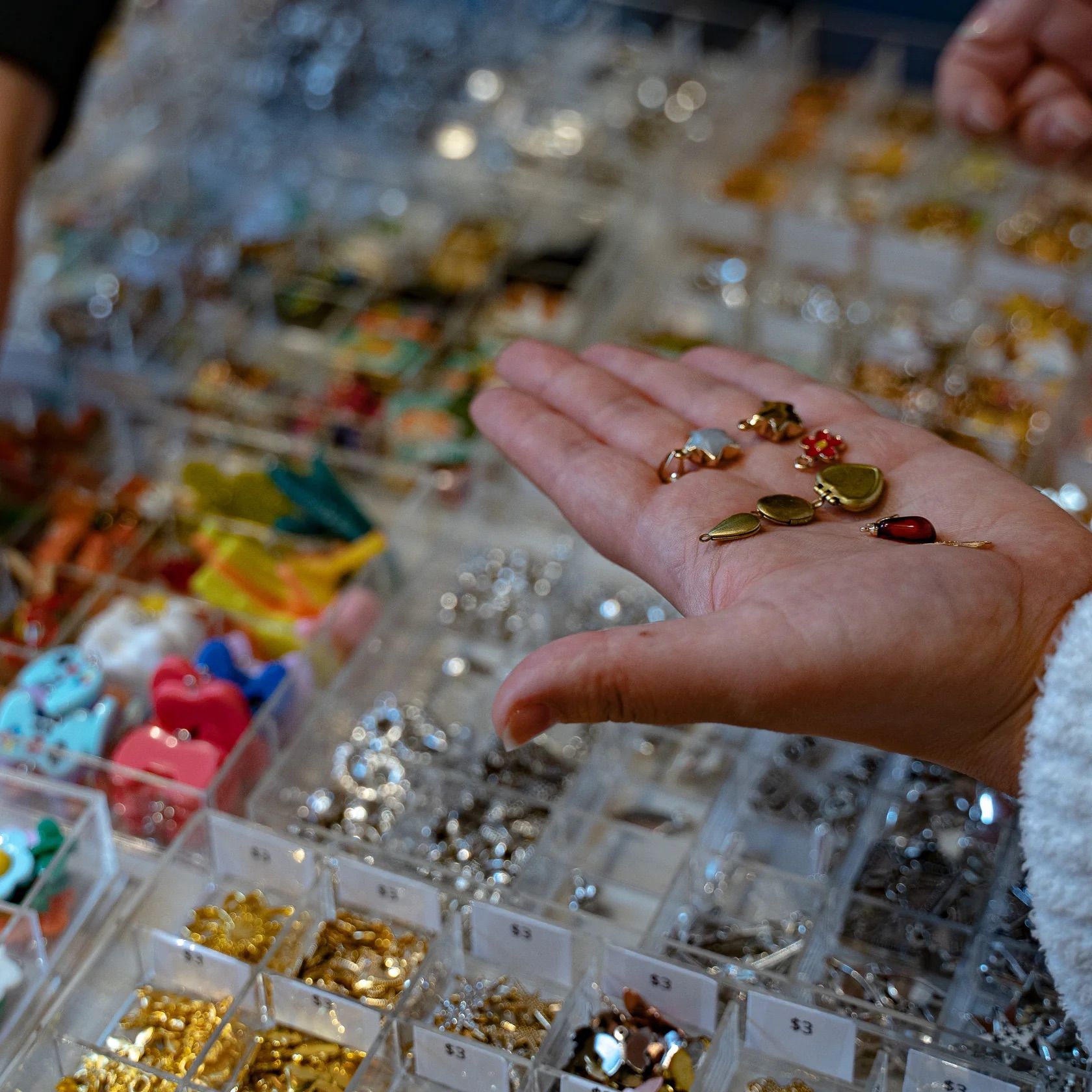 Open palm holding assorted small jewelry charms and pendants above clear organizer bins with colorful beads and gold and silver findings on a craft supply display