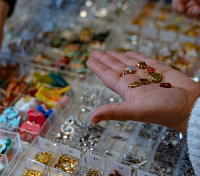 Open palm holding assorted small jewelry charms and pendants above clear organizer bins with colorful beads and gold and silver findings on a craft supply display