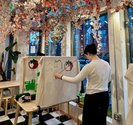 Person working on textile art on an upright canvas frame in a bright urban craft studio with hanging cherry blossom decorations, disco balls, tomato and green tufted designs, checkered floor and large windows overlooking a city street.