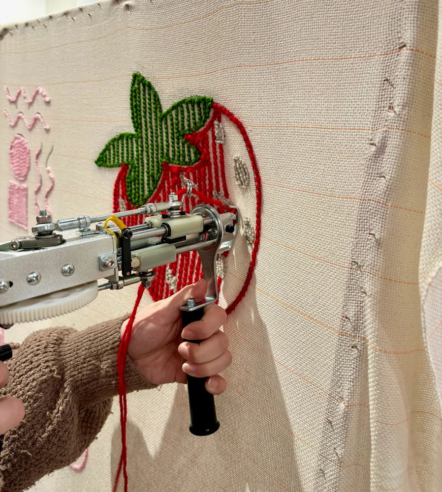 Close-up of artist's hands using a handheld tufting gun to punch red yarn into stretched canvas, forming a stitched strawberry with green leaves in a textile studio