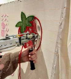 Close-up of artist's hands using a handheld tufting gun to punch red yarn into stretched canvas, forming a stitched strawberry with green leaves in a textile studio