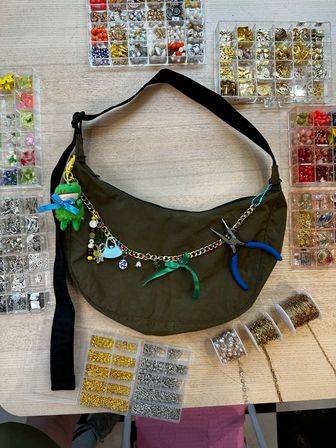 Overhead shot of an olive-green crescent shoulder bag on a light wood craft table, surrounded by clear organizers of colorful beads and charms, spools of gold and silver chain, blue-handled jewelry pliers, and a charm-decorated chain with a green plush keychain and ribbon.