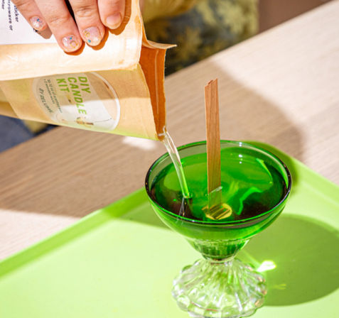 Hand pouring melted wax from a DIY candle kit pouch into a bright green glass dish fitted with a wooden wick on a light wood tabletop — close-up DIY candle-making scene.