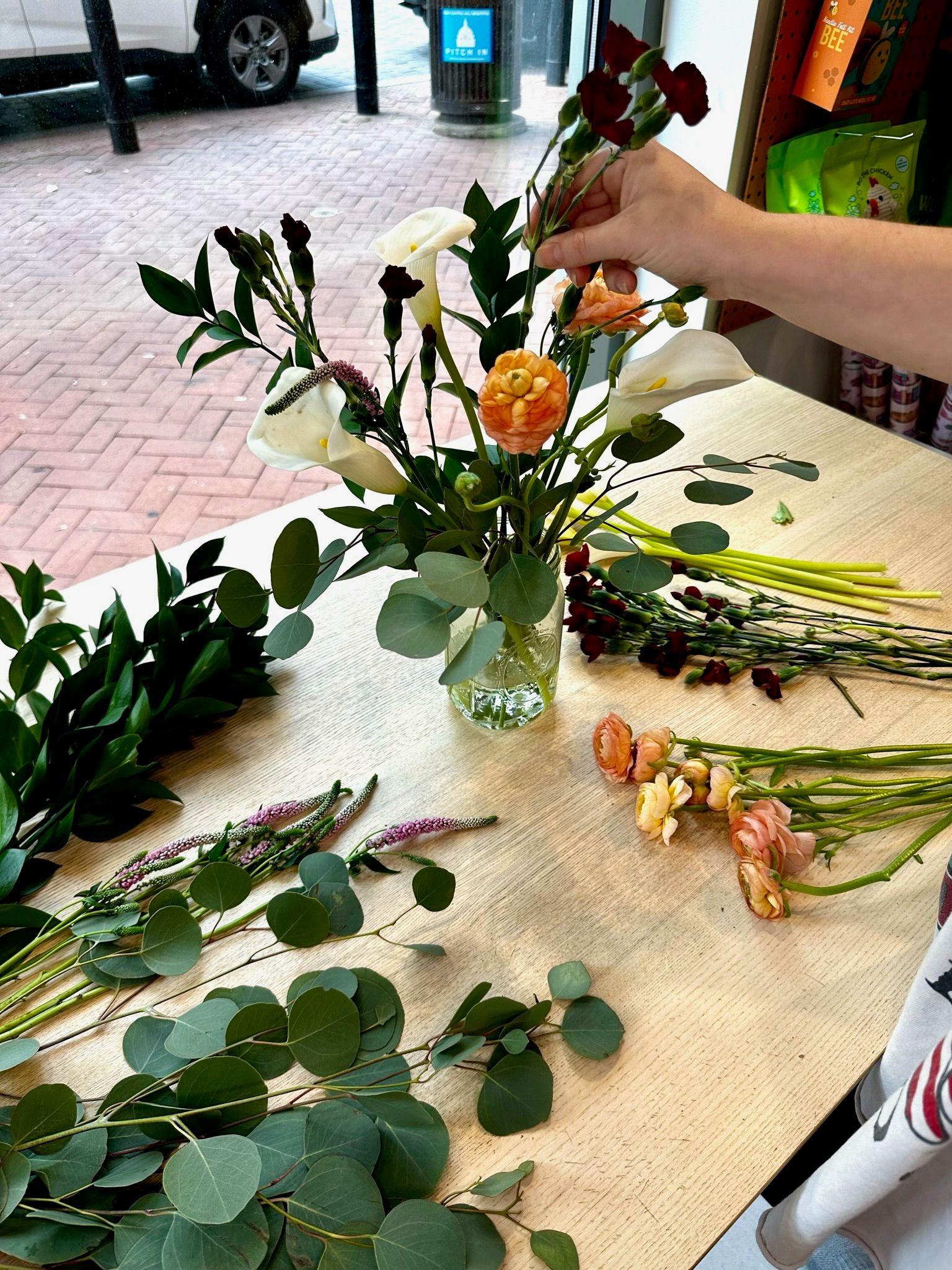 Hand arranging a spring bouquet in a glass vase on a shop table with white calla lilies, peach ranunculus, dark red carnations and eucalyptus alongside loose stems by a storefront window overlooking a brick sidewalk.