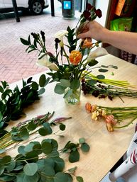 Hand arranging a spring bouquet in a glass vase on a shop table with white calla lilies, peach ranunculus, dark red carnations and eucalyptus alongside loose stems by a storefront window overlooking a brick sidewalk.
