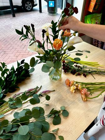 Hand arranging a spring bouquet in a glass vase on a shop table with white calla lilies, peach ranunculus, dark red carnations and eucalyptus alongside loose stems by a storefront window overlooking a brick sidewalk.