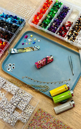 DIY jewelry workspace with compartment boxes of rainbow and letter beads, rhinestones, chains, tweezers and colorful lighters arranged on a blue tray